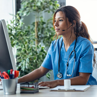 Woman in light blue scrubs sits in front of a computer.